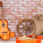an autumn harvest arrangement against a brick wall, including an acoustic guitar, apples, wheat, pumpkins and a pair of well-worn boots.